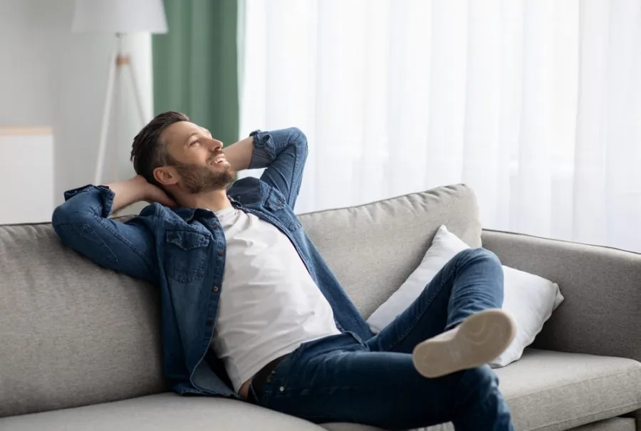 A man relaxes on a grey sofa, smiling contentedly in a bright living room.